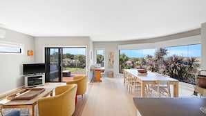 A modern living room with a flat-screen TV, a dining area with a large table and chairs, and a balcony with a view of the surrounding landscape. - Beautiful spacious pet friendly home next to the ocean (Smiths Beach)