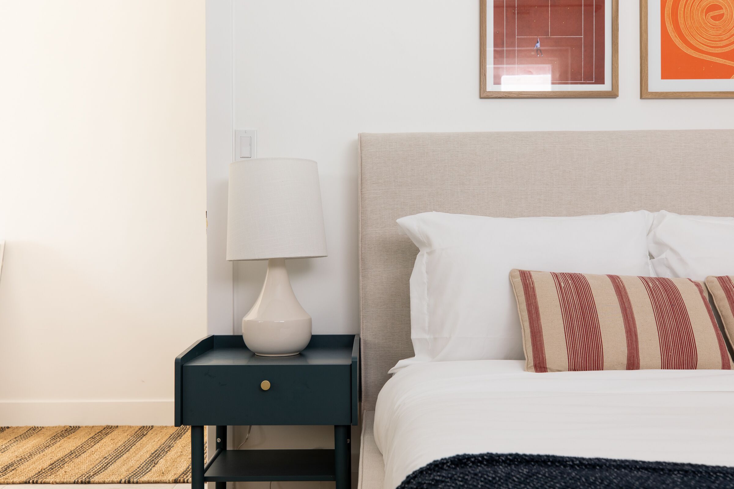 Una cama bien hecha con almohadas blancas y a rayas, una lámpara de noche sobre una mesita oscura y un cuadro en la pared.