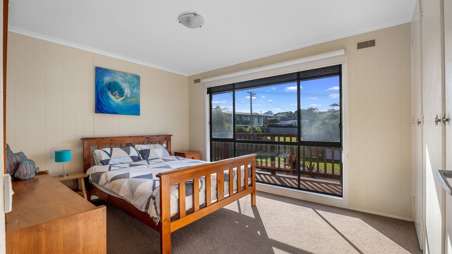 A bedroom with a bed, a nightstand, a lamp, a painting, and a balcony with a view of the neighborhood.