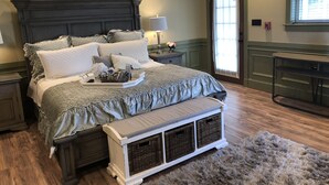 A neatly arranged bedroom with a large bed, bedside tables, a nightstand, a bench with baskets, and a window with blinds. - Guest Quarters at the Inn - 103 (North Beach)
