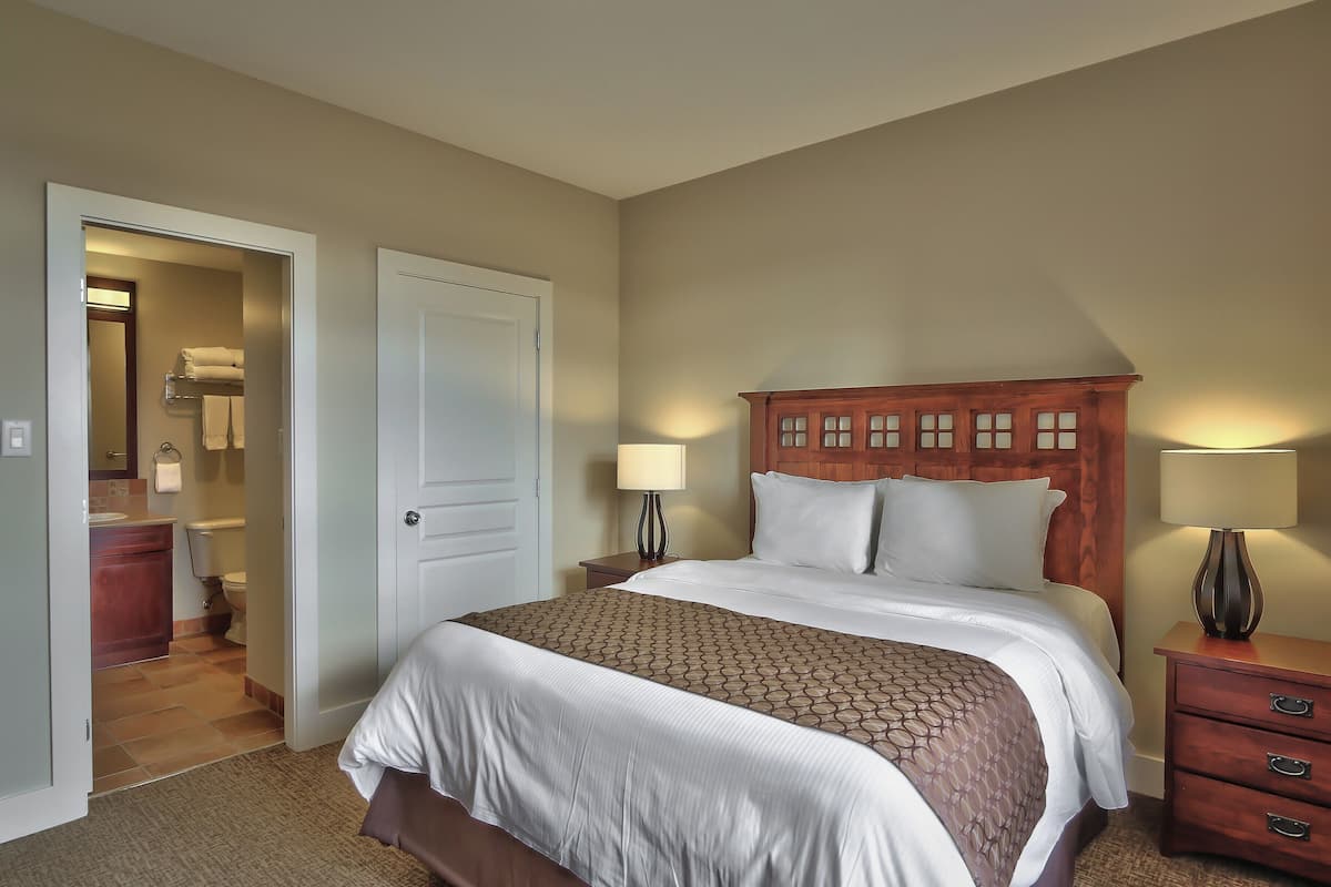 A hotel room with a bed, a nightstand with a lamp, a wooden headboard, and a door leading to a bathroom.