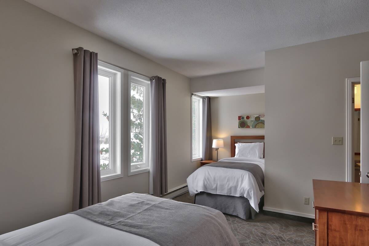 A hotel room with two beds, a nightstand, a lamp, and a view of trees through the windows.
