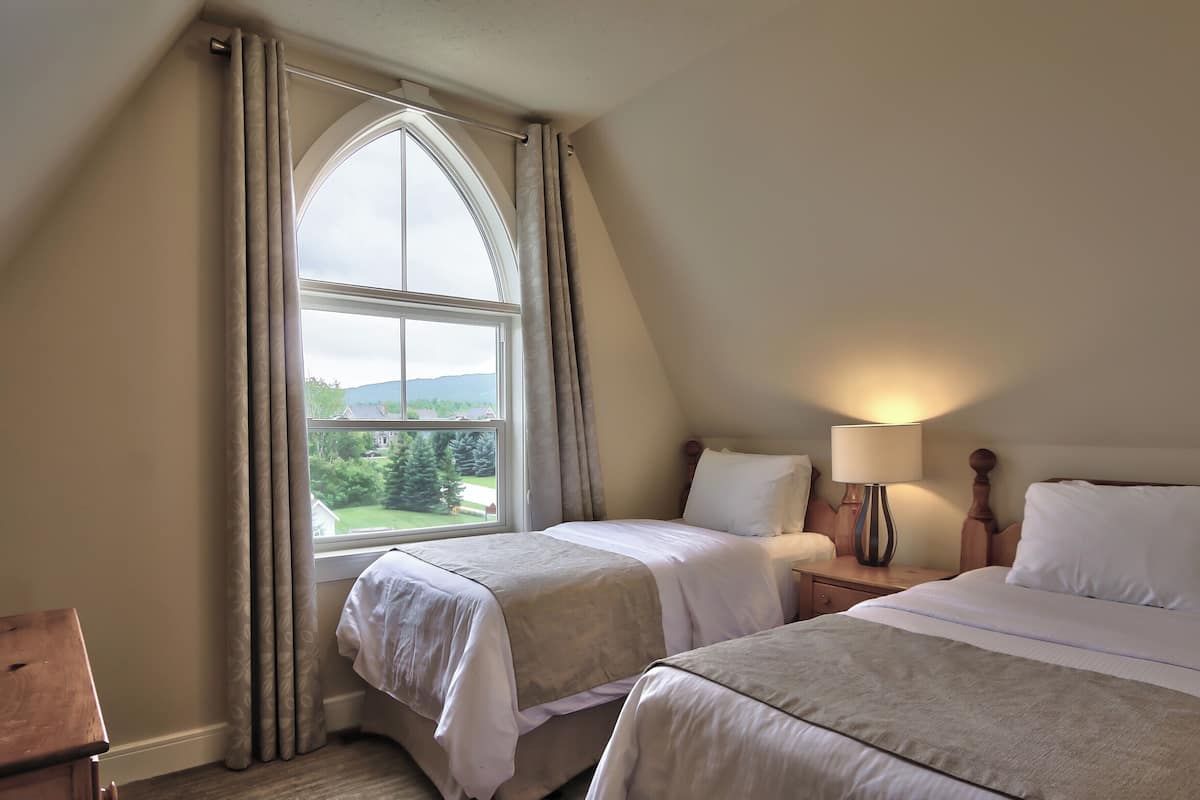 A bedroom with two beds, a window with curtains, a bedside table with a lamp, and a view of the outdoors.