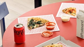 A table with a pizza box, a can of Coca-Cola, a small container of sauce, and a bottle of water.