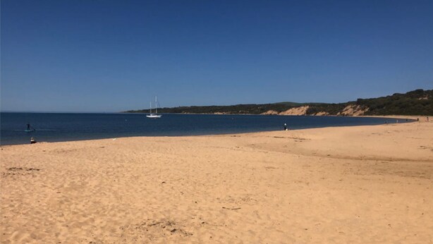 En bred sandstrand med en klarblå himmel, en segelbåt i fjärran och ett kuperat landskap i bakgrunden.