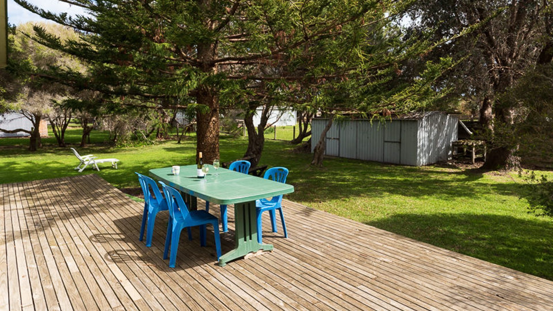 An outdoor wooden deck with a table and chairs, surrounded by trees and a shed.