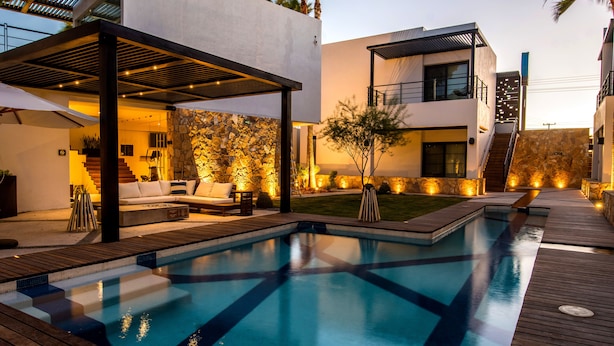 A modern outdoor pool area with a wooden deck, a stone wall, and a covered seating area. - Marsella 45 (Hermosillo)