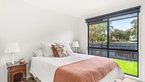A bedroom with a large window, a bed with white bedding, two bedside lamps, and a small wooden nightstand with a vase and a heart-shaped ornament. - Perfect family getaway, walk to beach and boat ramp. (Cowes)