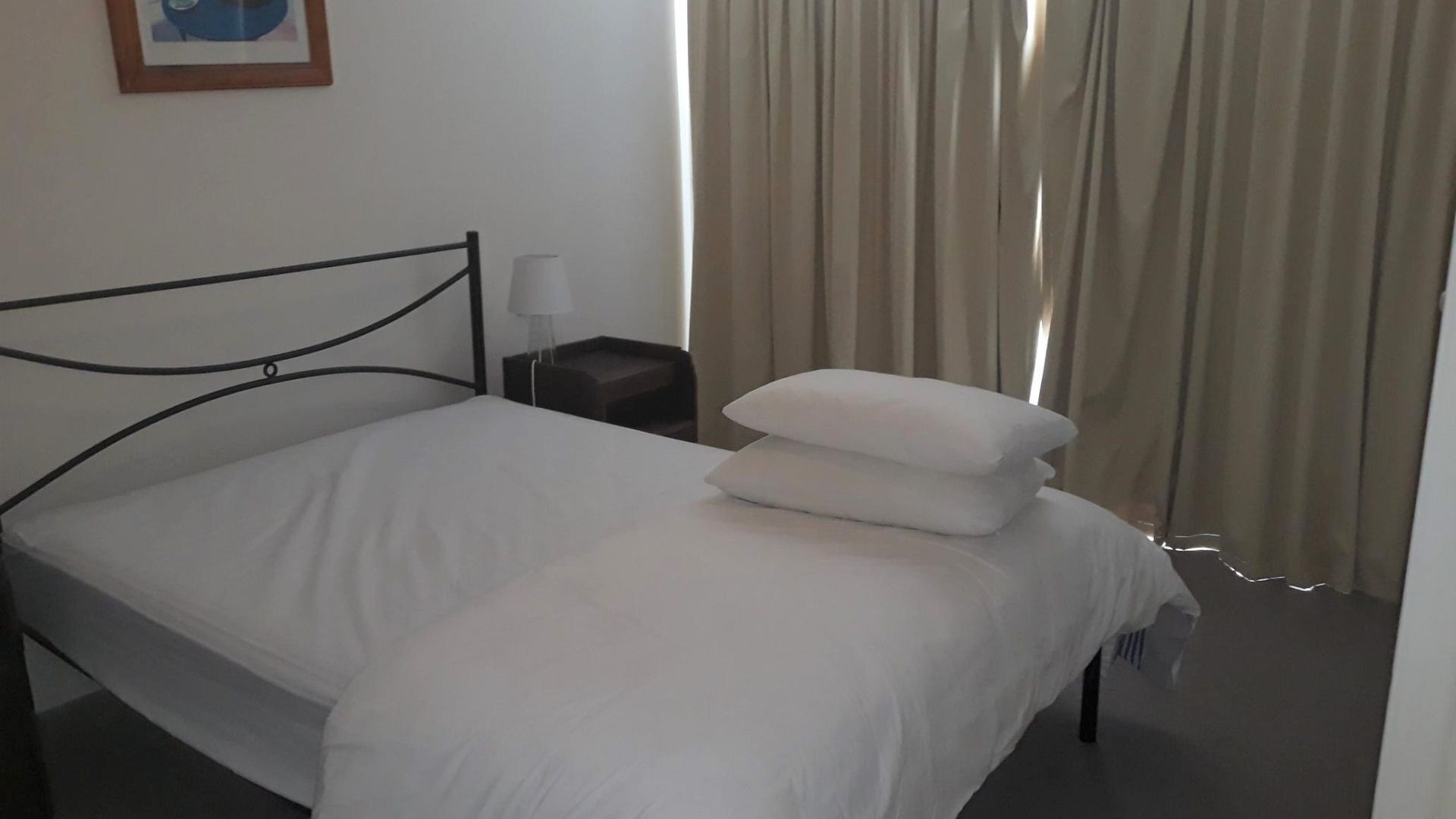 A neatly made bed with white linens in a simple room with a metal headboard, a small bedside table with a lamp, and beige curtains.