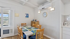 Un coin repas avec une table blanche, des chaises bleues et une nappe à motifs. Il y a une horloge au mur et une fenêtre donnant sur un paysage verdoyant.