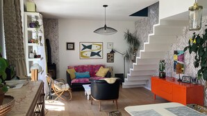 En moderne stue med trappe, en sofa med farverige puder, et spisebord og en reol.