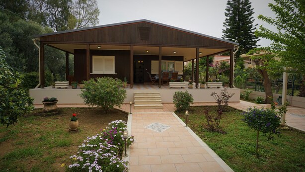 Una casa moderna con un porche cubierto, un jardín con flores y arbustos, y un camino pavimentado que conduce a la entrada.