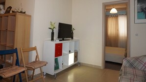Una sala de estar acogedora con un televisor sobre un mueble blanco, una silla de madera, un sofá y una estantería.