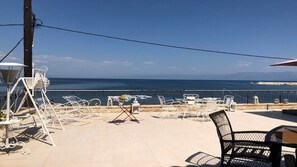 Ein Sitzbereich im Freien mit Metallstühlen und Tischen, mit Blick auf die Küste, das Meer und ferne Berge.