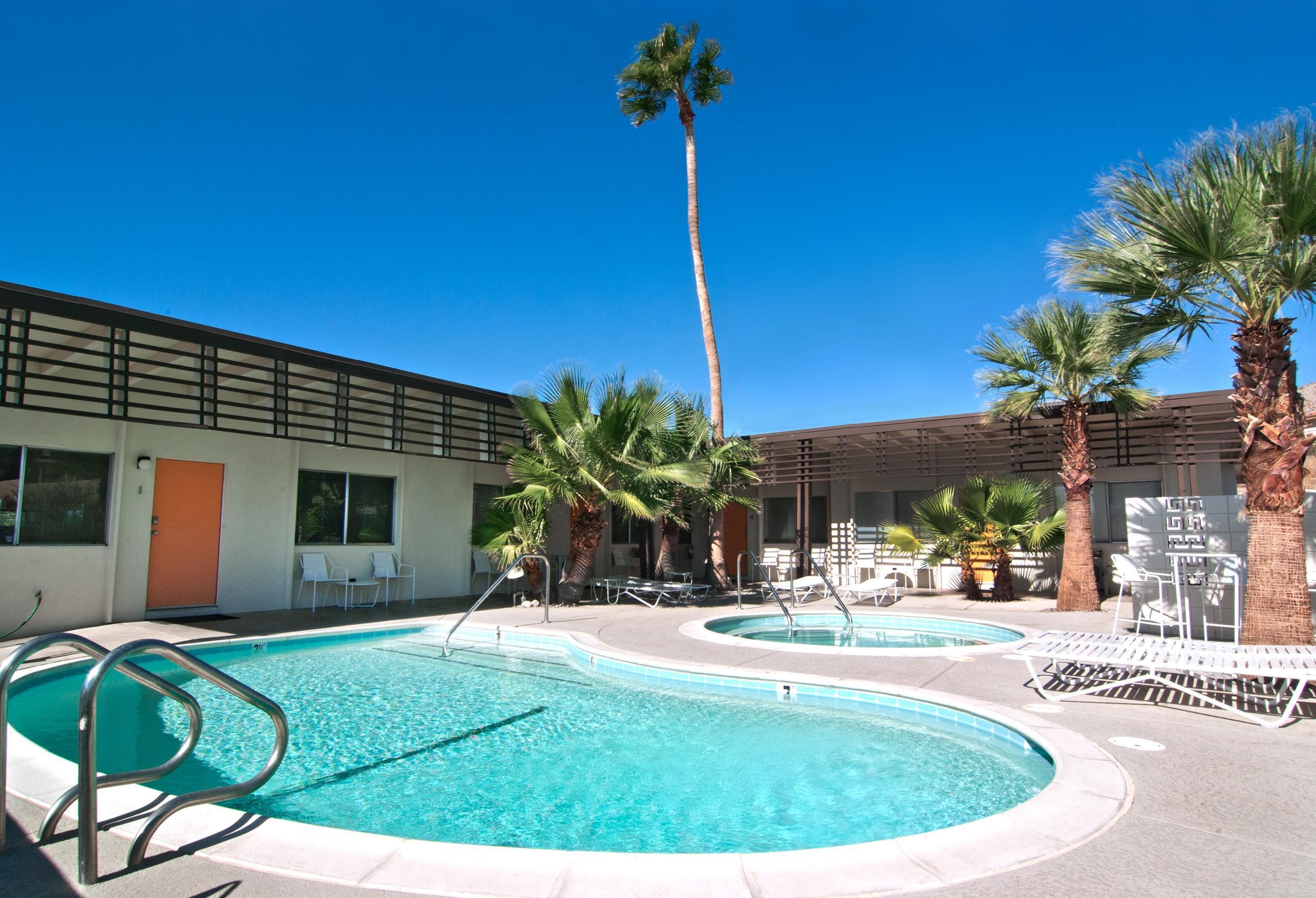 Un espace piscine comprenant deux piscines, des palmiers, des chaises longues et un bâtiment doté d’une porte et de fenêtres.