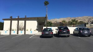 A parking lot with three cars parked in front of a building with a sloped roof and palm trees. - The Getaway Studio #3: Pool, Mountain View, Kids welcome (Desert Hot Springs)