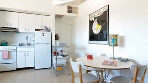 A modern kitchen with white cabinets, a stove, and a refrigerator. There is a dining table with chairs and a framed picture on the wall. - The Getaway Studio #3: Pool, Mountain View, Kids welcome (Desert Hot Springs)