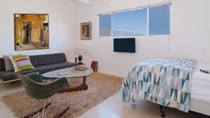 A modern room with a sofa, a glass coffee table, a bed with a patterned bedspread, a painting, and a TV on the wall. - The Getaway Studio #3: Pool, Mountain View, Kids welcome (Desert Hot Springs)