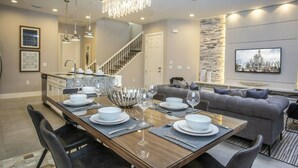 A modern dining area with a long wooden table set for a meal, a staircase, and a living area with a gray sofa and a television.