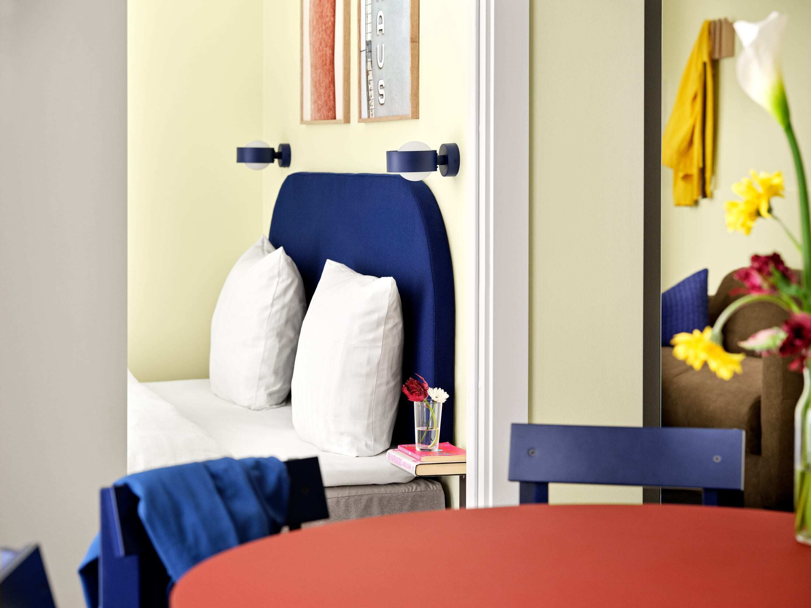 A room with a bed, a table, and a chair. There is a vase with flowers on the table.