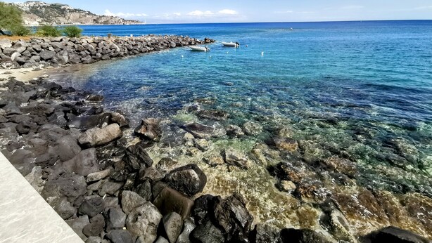 A rocky shoreline with clear water, a boat, and a distant lighthouse.