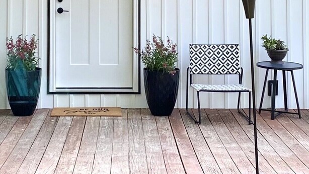 A small, white wooden building with a sign reading "Rider's Den." It features a door with a window, two potted plants, a patterned chair, and a small table with a plant. - Private cottage located on The Carriage House Inn Estate (Mena)