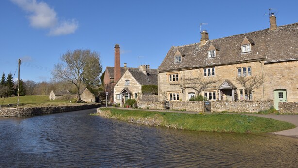 A stone building with multiple windows and a chimney, situated beside a calm waterway. - Period cottage in idyllic riverside location in prime Cotswolds village (Lower Slaughter)