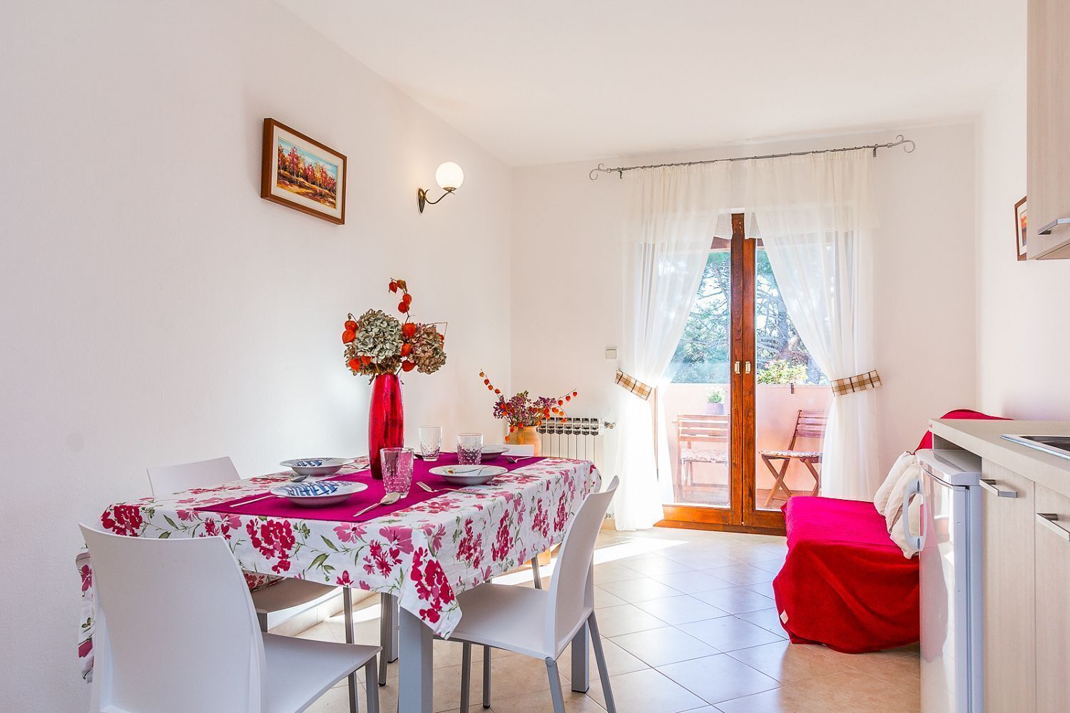 A dining area with a table set for four, a red vase with flowers, and a view of the outside through a glass door.