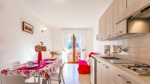 A modern kitchen with light wood cabinets, a built-in oven, and a sink. There is a dining area with a table set for two, chairs, and a vase with flowers.