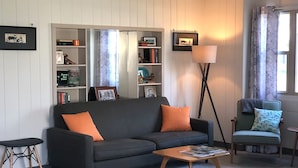 A living room with a dark grey sofa, an orange cushion, a wooden coffee table, a floor lamp, a bookshelf, and a chair with a blue cushion. - Blue Steel Lustron - Rare - Near Starved Rock Country (Grand Ridge)
