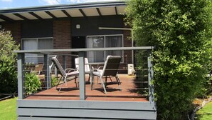 A deck with outdoor furniture, a brick building in the background, and a well-maintained garden. - Honeysuckle Beach Cottage - Silverleaves (SILVERLEAVES)