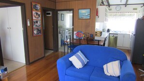 A living room with a blue sofa, wooden flooring, a dining area with a table and chairs, a kitchen with appliances, and a door leading to another room. - Honeysuckle Beach Cottage - Silverleaves (SILVERLEAVES)
