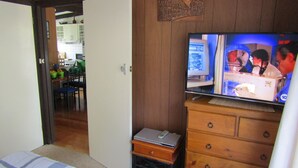 A living room with a television on a wooden cabinet, a laptop on a small table, and a kitchen in the background. - Honeysuckle Beach Cottage - Silverleaves (SILVERLEAVES)