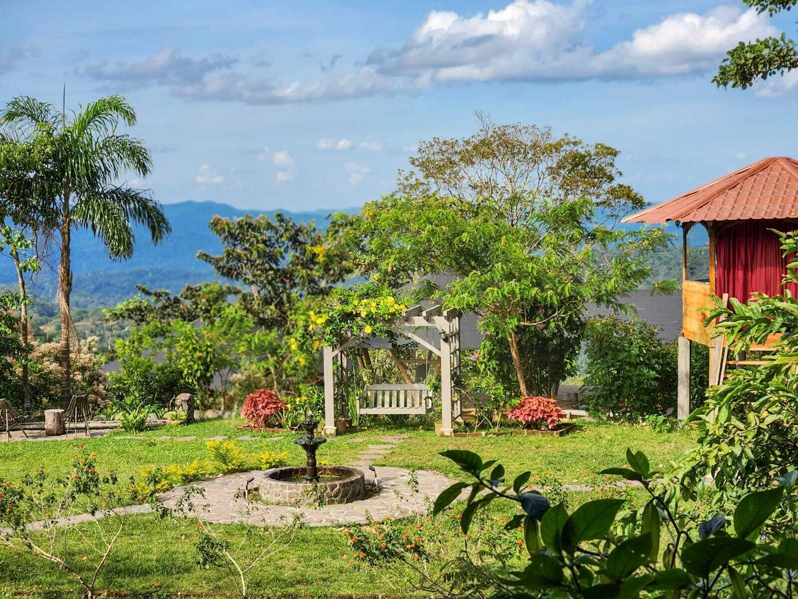 A lush garden with a fountain, a gazebo, and a building with a red roof.