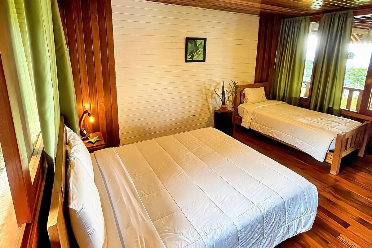 A room with two beds, wooden flooring, a small bedside table with a lamp, and a framed picture on the wall.