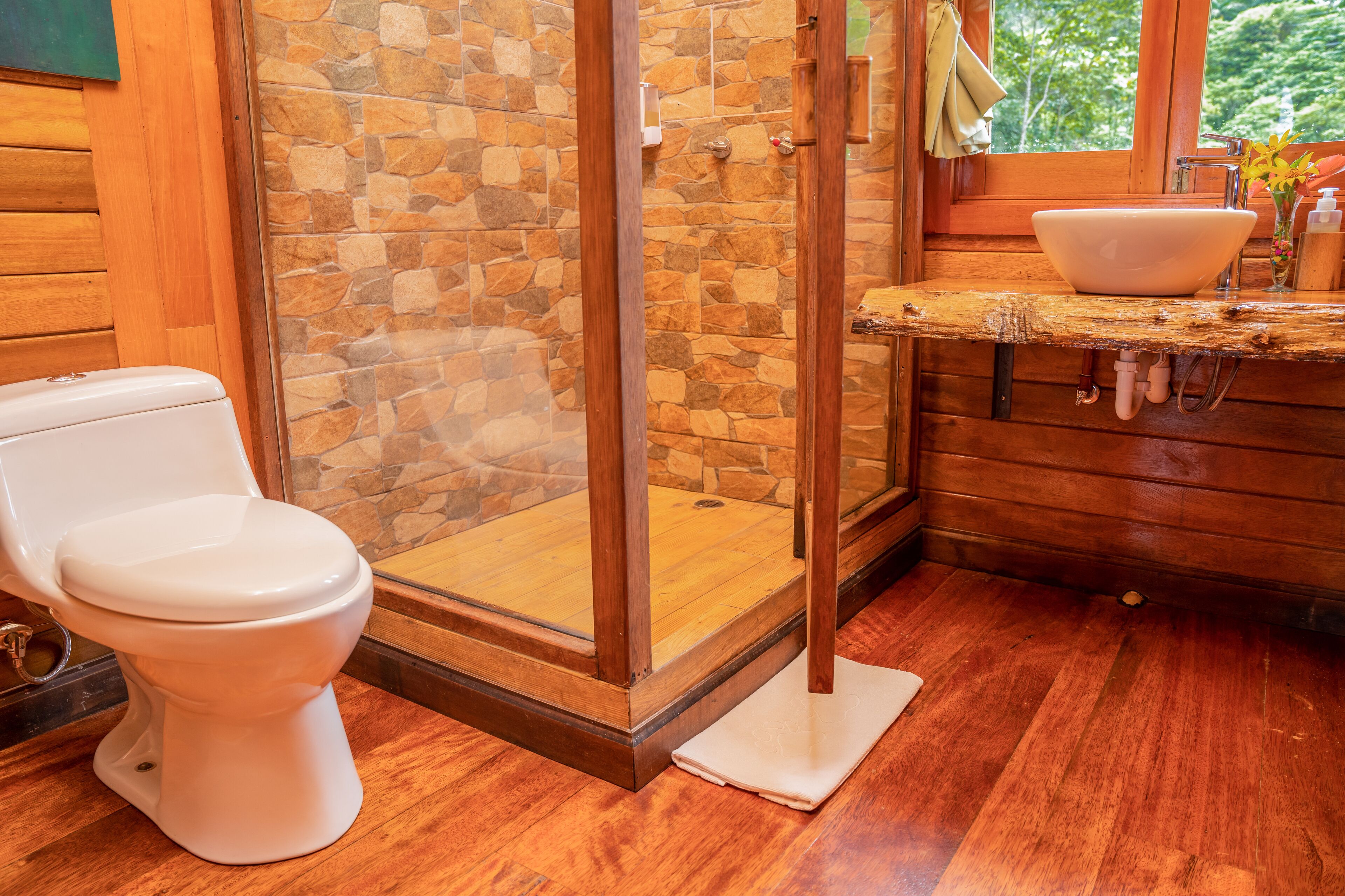 Un baño de madera con cabina de ducha de vidrio, un inodoro blanco y un tocador de madera con lavabo.