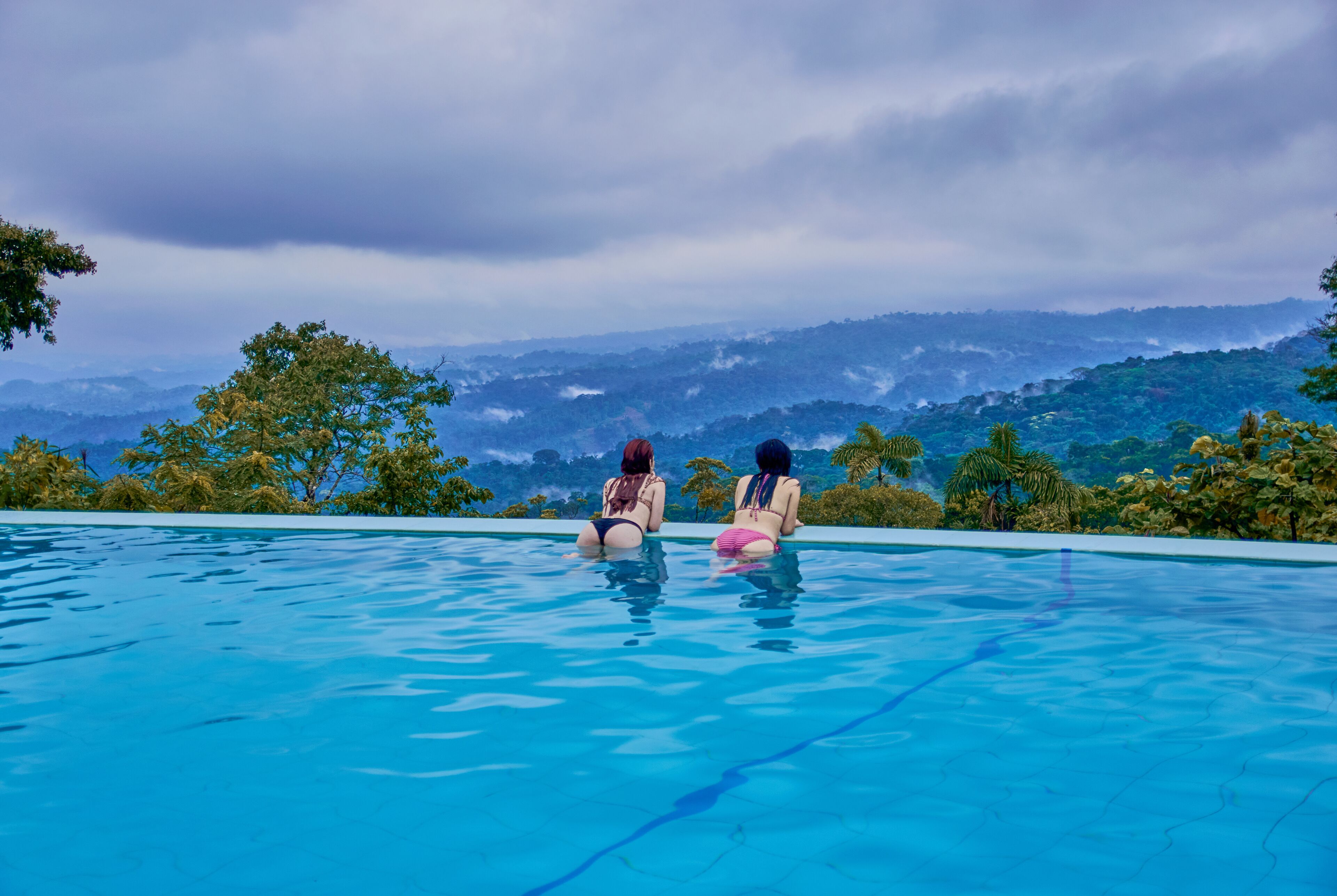 A pool with two people sitting at the edge, overlooking a mountainous landscape.