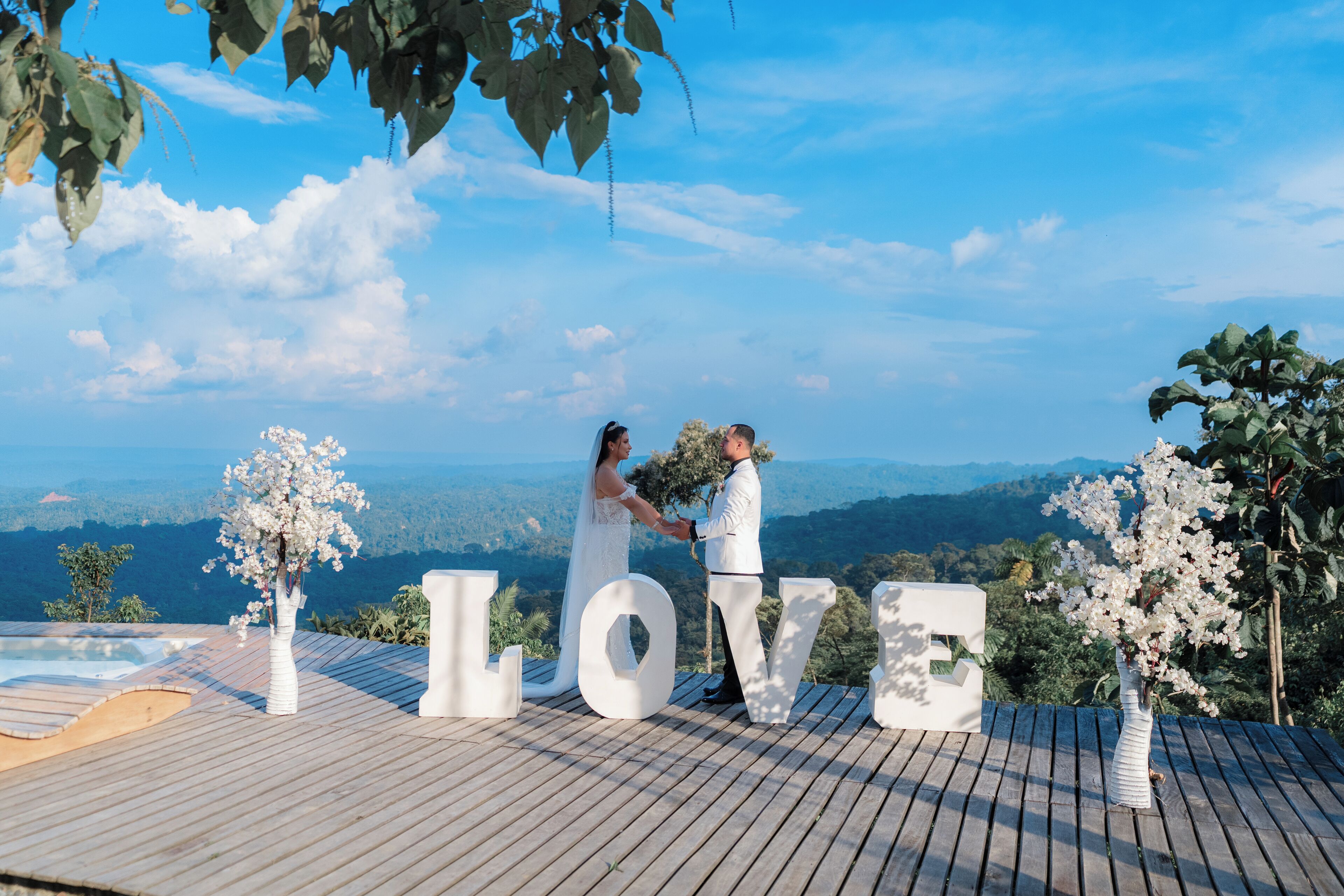 A couple standing on a wooden deck with large white letters spelling "LOVE" and a view of mountains in the background.