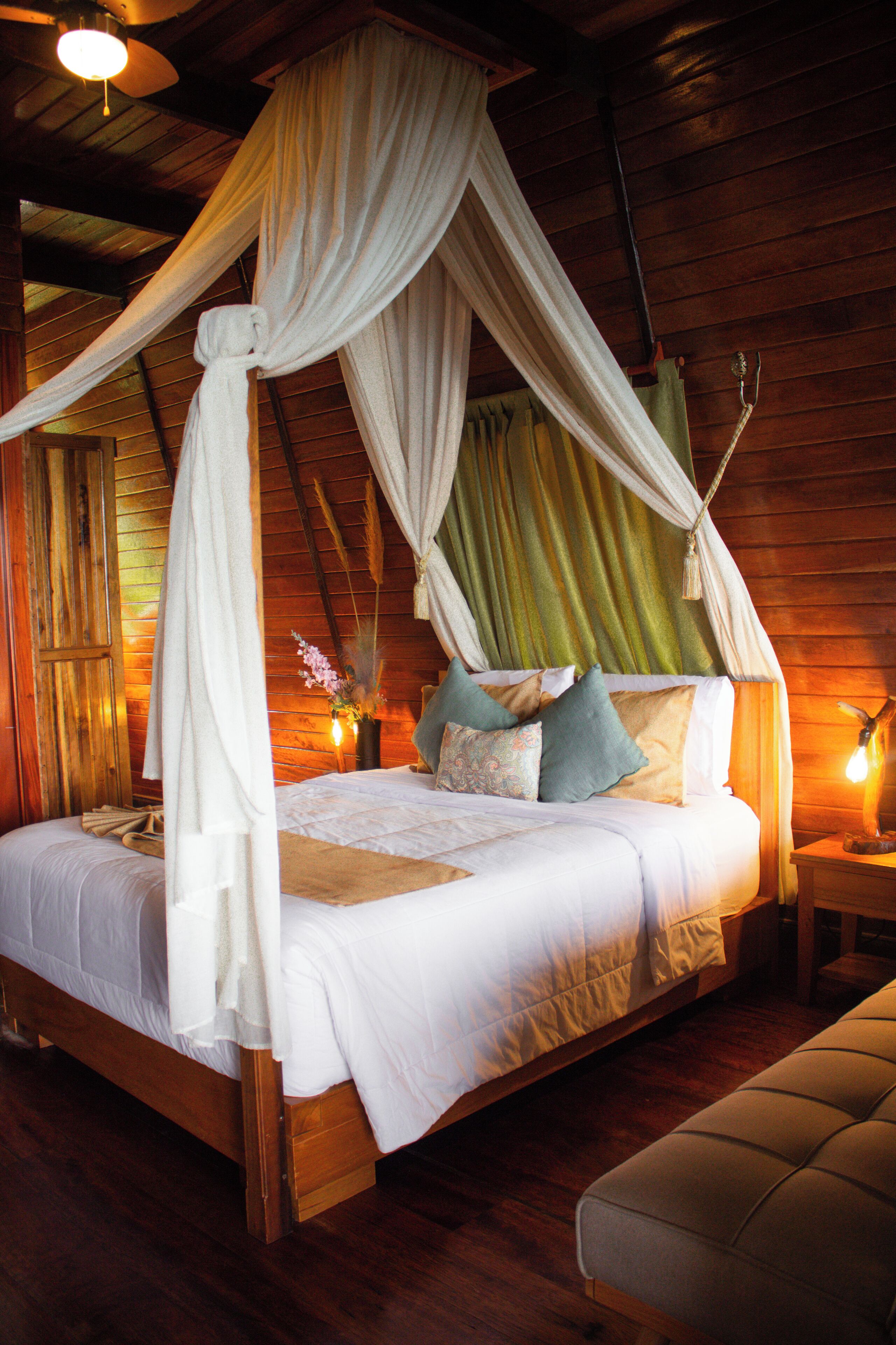 A wooden room with a four-poster bed, a bench, and a nightstand with a lamp.