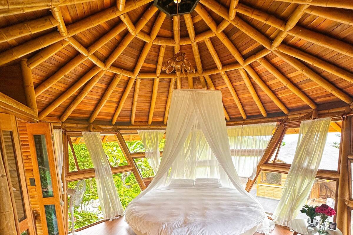 A circular bed with a canopy in a room with a wooden ceiling and large windows.