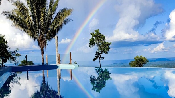 A clear pool with reflections of palm trees and a rainbow, surrounded by tropical vegetation and a scenic view.