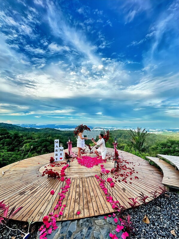 A wooden deck with a view of a mountainous landscape, a sign reading "MARRY ME?", and scattered rose petals.
