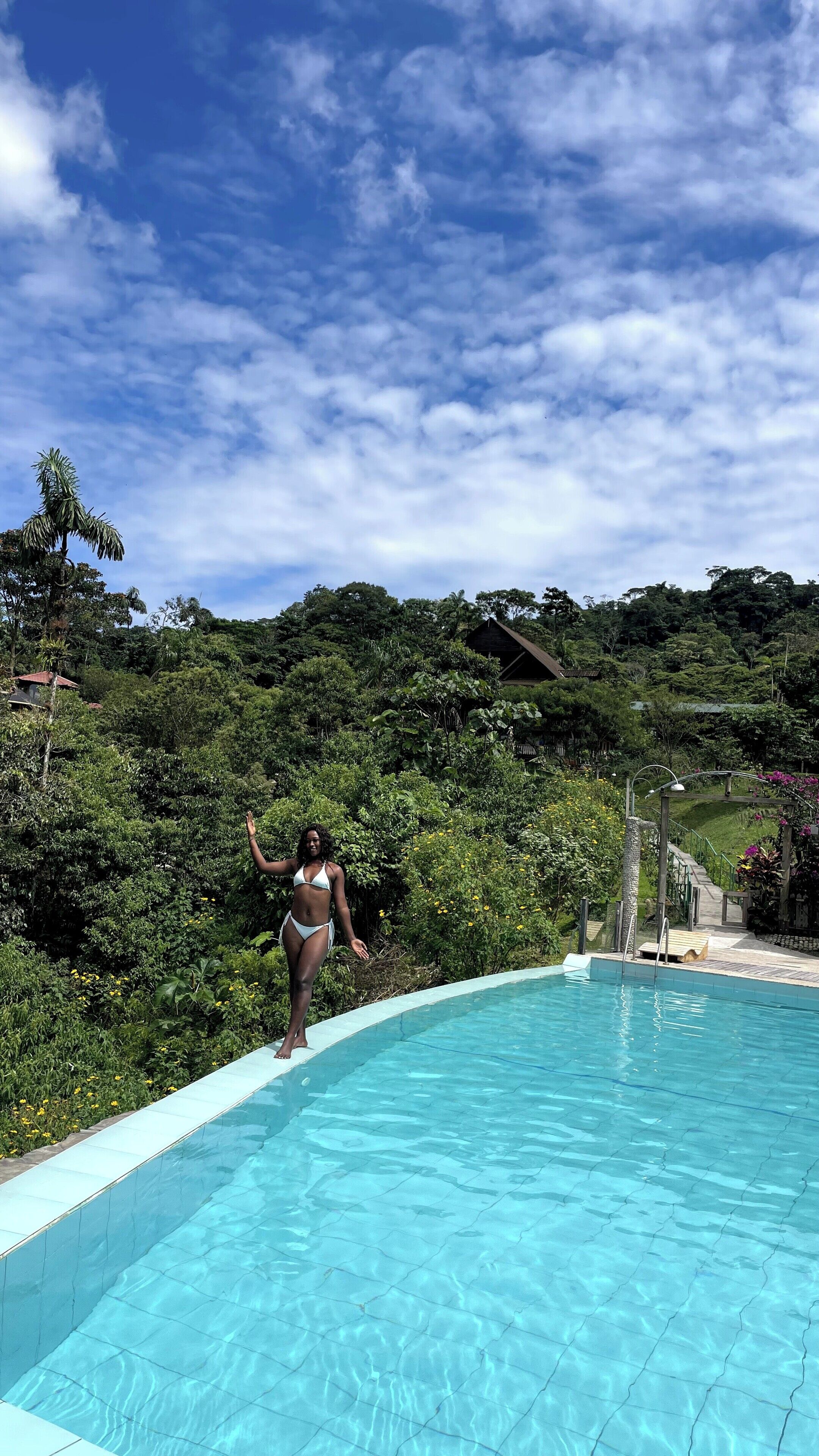 A woman in a bikini standing at the edge of a clear blue pool with lush greenery and a wooden deck in the background.