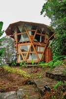 Una estructura de madera con diseño geométrico, rodeada de abundante follaje y un pequeño arroyo.