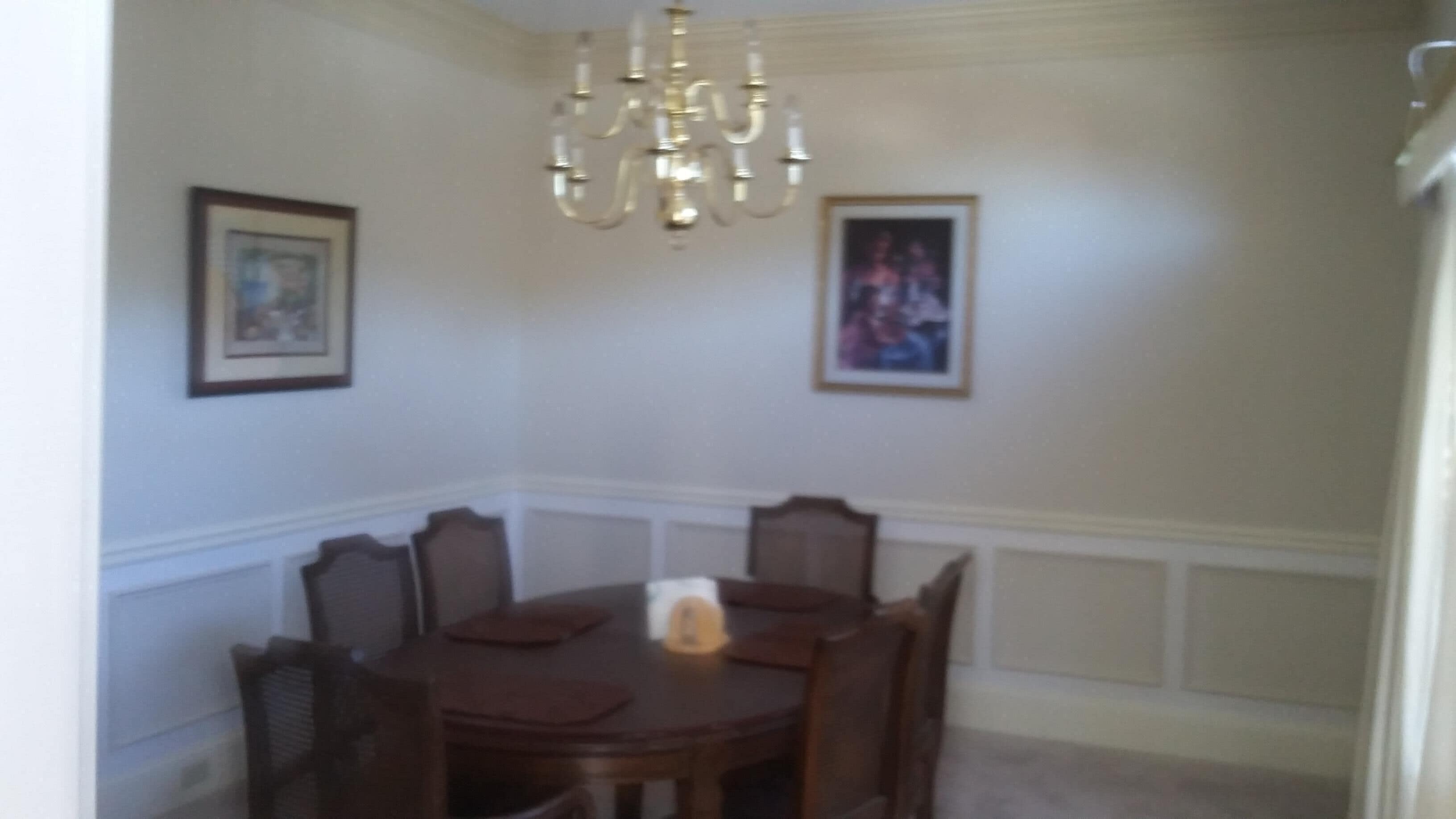 Un comedor con una mesa redonda, sillas, una araña y dos cuadros en las paredes.