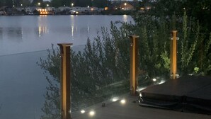 A wooden deck with railings and lights, overlooking a calm body of water with distant buildings. - Beautiful Lakeside lodge with Hot Tub (LINCON)