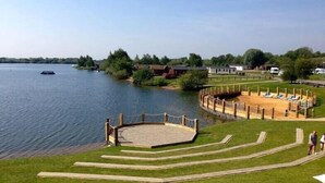 A lakeside area with a wooden deck, a sandy play area, and a boat on the water. - Beautiful Lakeside lodge with Hot Tub (LINCON)