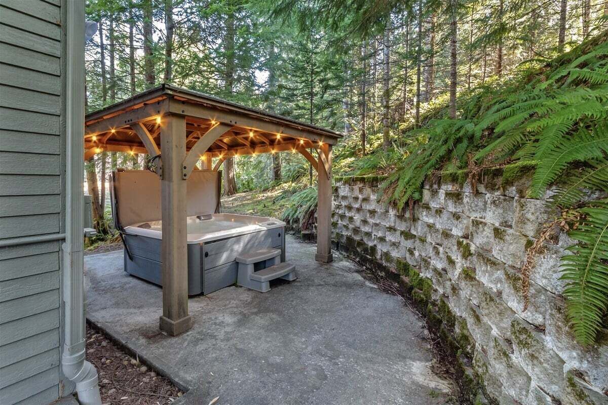 Een hottub onder een houten pergola met lichtslingers, omringd door een stenen muur en weelderige begroeiing.