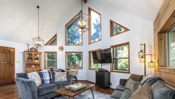 A spacious living room with a grey sofa, a wooden coffee table, and a television. Large windows allow natural light and views of the outdoors. - Cedar Grove Lodge | BBQ | Wifi, Hot tub, AC (Packwood)