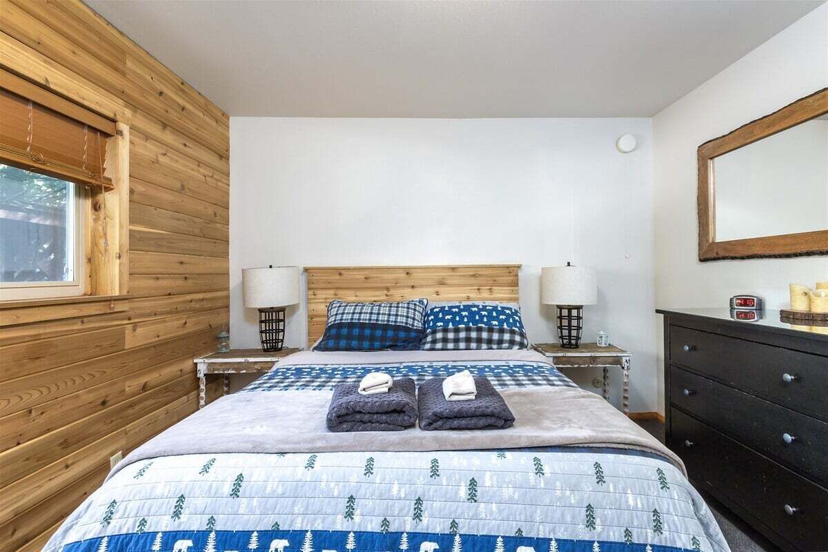 A bedroom with a wooden headboard, a bed with a blue and white patterned bedspread, two pillows, a nightstand with a lamp, and a dresser with a mirror.